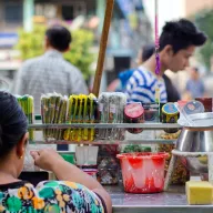 Woman at a betel stall with a lime pot and a splattered bucket, customers waiting in the background