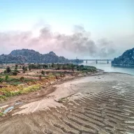 Sandy riverbank with ripple patterns, smoke clouds and a bridge, from above