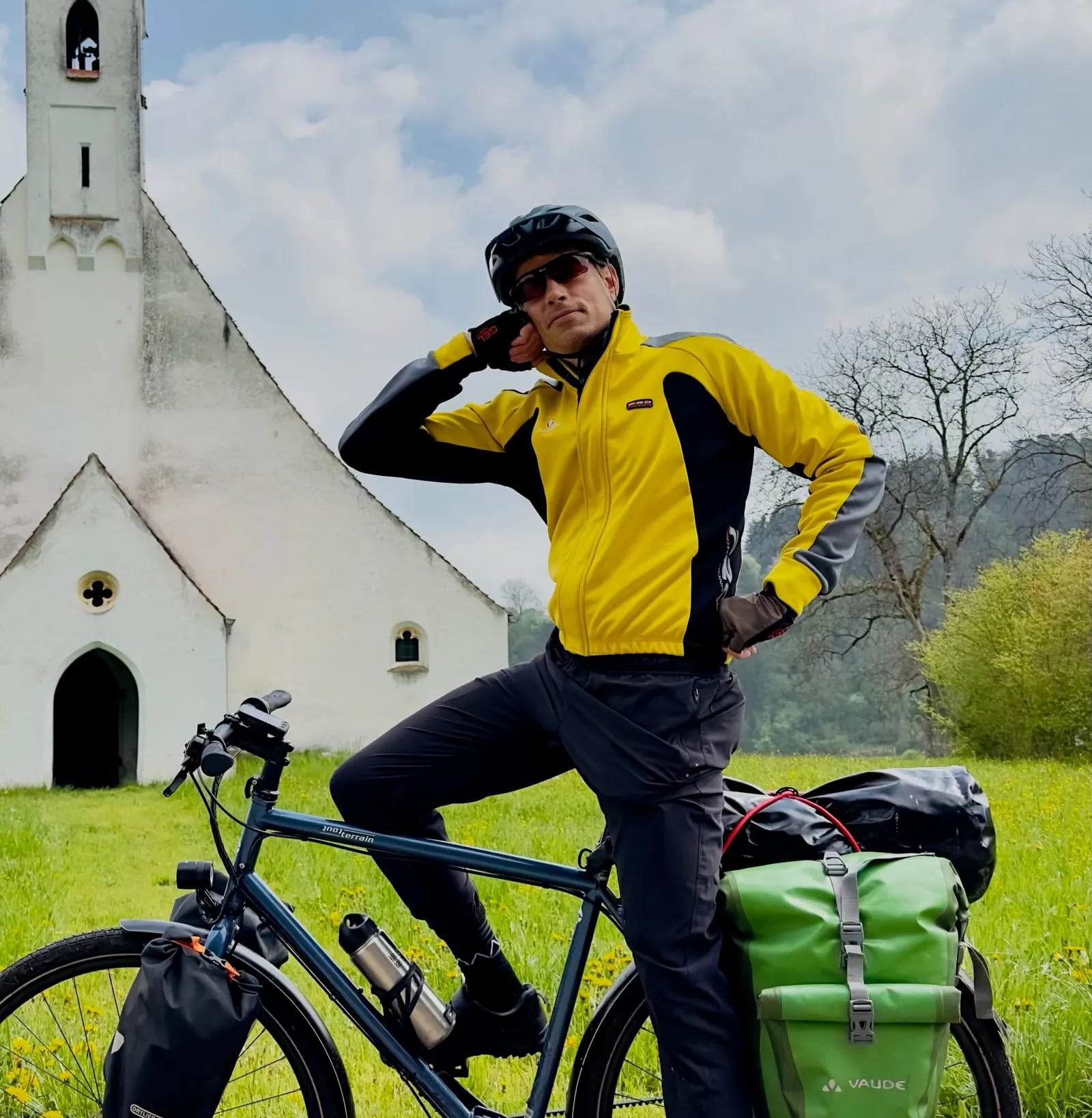 Samuel Mellert mit Helm und Sonnenbrille auf seinem Reiserad mit Packtaschen, dahinter eine weiße Kirche