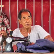 Elderly man at a sewing machine in front of a red wall