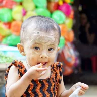 Toddler with thanaka eating, colourful balloons behind